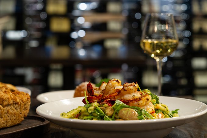 Pasta dish served on a white plate on a wooden table with a glass of white wine and wine wall background. Loaf of bread also on the table.